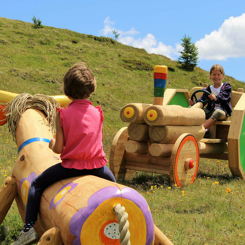 Kindermeile Golzentipp Obertilliach Zwei Mädchen, die auf einer bunten Holzkuh bzw. auf einem bunten Holztraktor sitzen. Dahinter ein grasbewachsener Berghang mit spärlichem Bewuchs und einem leicht bewölkten Himmel.