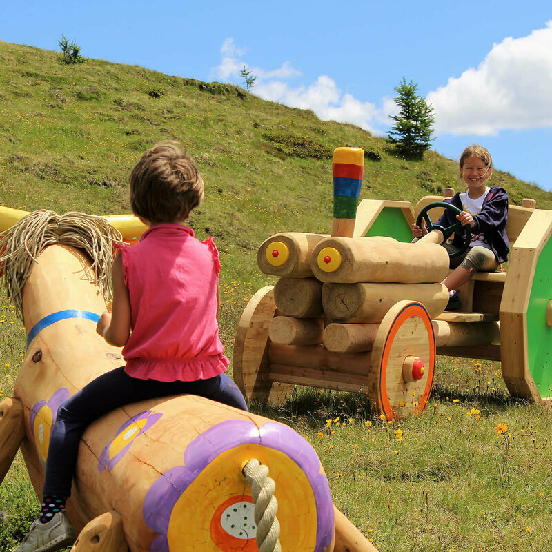 Zwei Mädchen, die auf einer bunten Holzkuh bzw. auf einem bunten Holztraktor sitzen. Dahinter ein grasbewachsener Berghang mit spärlichem Bewuchs und einem leicht bewölkten Himmel.