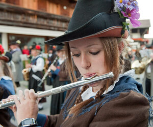 Ein Mädchen in Tracht spielt Querflöte. Im Hintergrund sind noch weitere Musikanten.