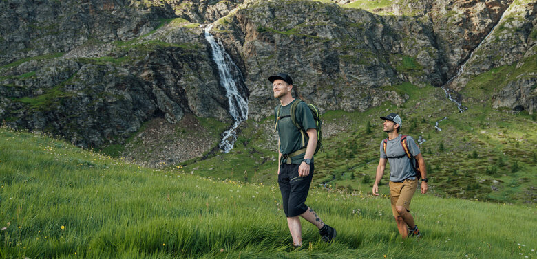 Passo dopo passo nella natura dell'Osttirol. Sono le esperienze a rimanere impresse. Zwei Männer wandern in Osttirols Bergen hinauf zum Geigensee durch hohes Bergwiesengras. Im Hintergrund ist ein kleiner Wasserfall zu sehen, der talwärts rinnt.