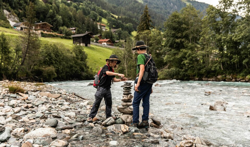 Zwei Kinder in Wanderkleidung und Hüten bauen am Ufer der Isel ein Stoanamandl.