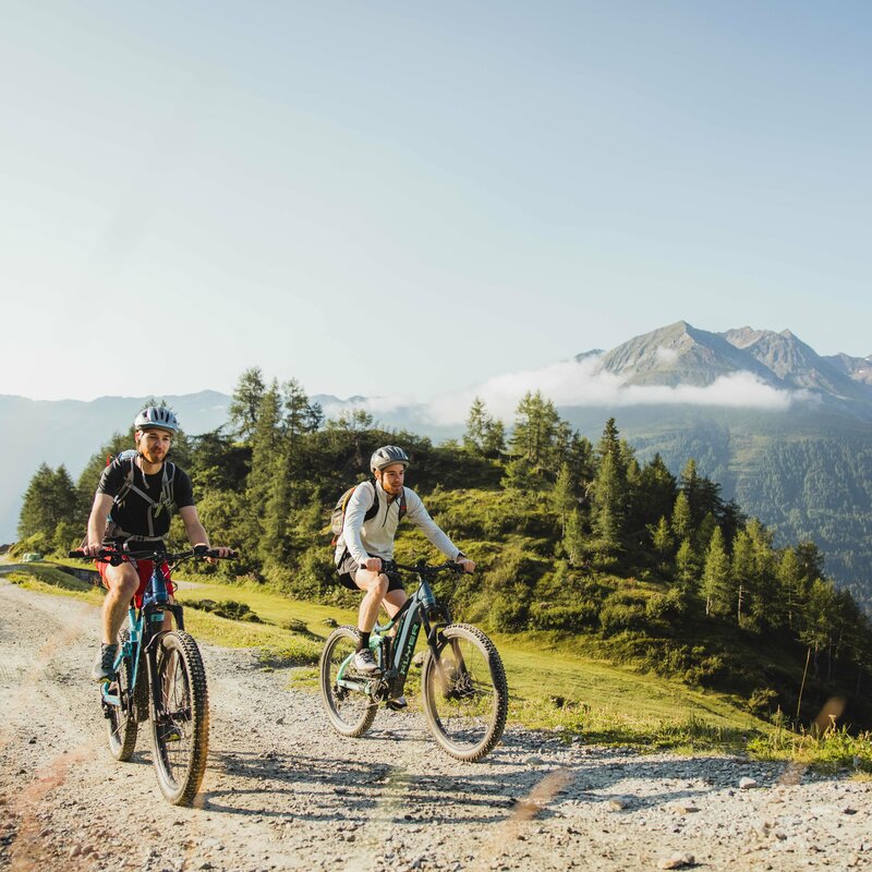 Biketour Timmeltal Praegraten Zwei junge E-Biker auf blau-grauen Bikes die bergwärts auf einem leicht ansteigenden Schotterweg fahren. Die Sonne scheint ihnen in den Rücken. Den Hintergrund bildet eine kleine, leicht bewaldete Erhebung hinter dieser es tief ins Tal zu gehen scheint. Ganz im Hinergrund erheben sich zwei Berge, deren Gipfelbereich eine dünne Nebelbank ziert.
