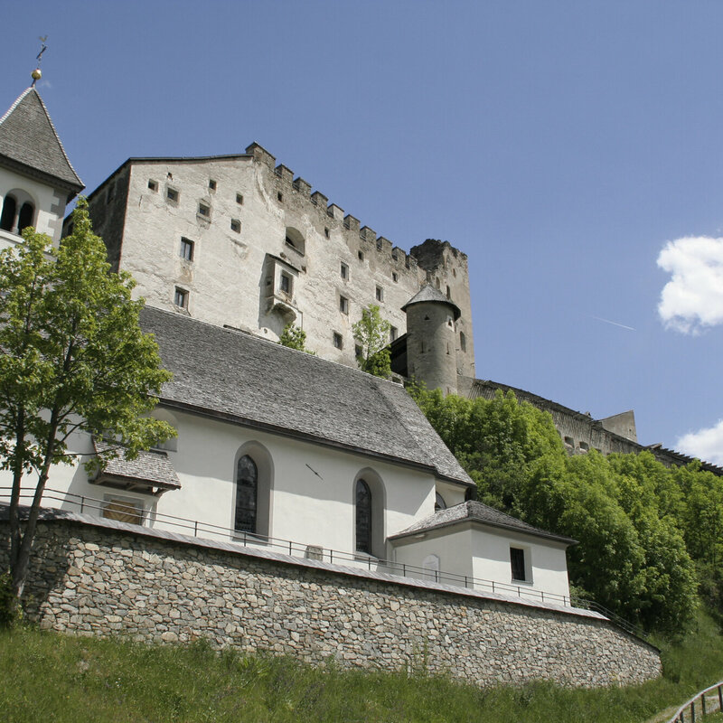 Die Burg Heinfels thront im Sommerlicht über der Landschaft, am Hang darunter liegt malerisch die kleine Kapelle.