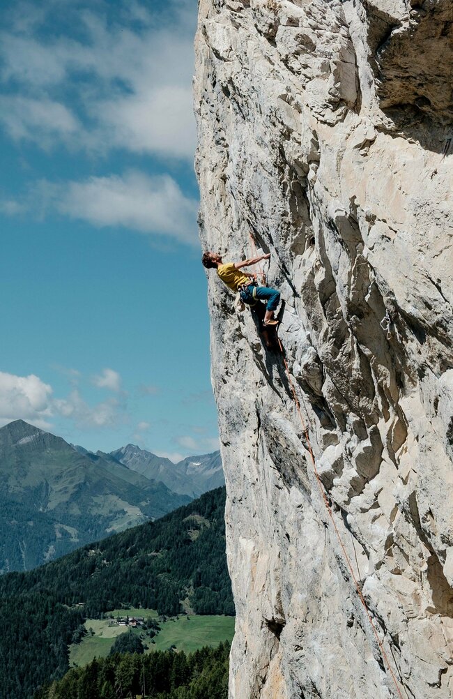 Ein Kletterer mit gelbem T-Shirt hängt an einer steilen Felswand im Klettergarten Falkenstein. Im Hintergrund erstreckt sich eine weitläufige Berglandschaft.