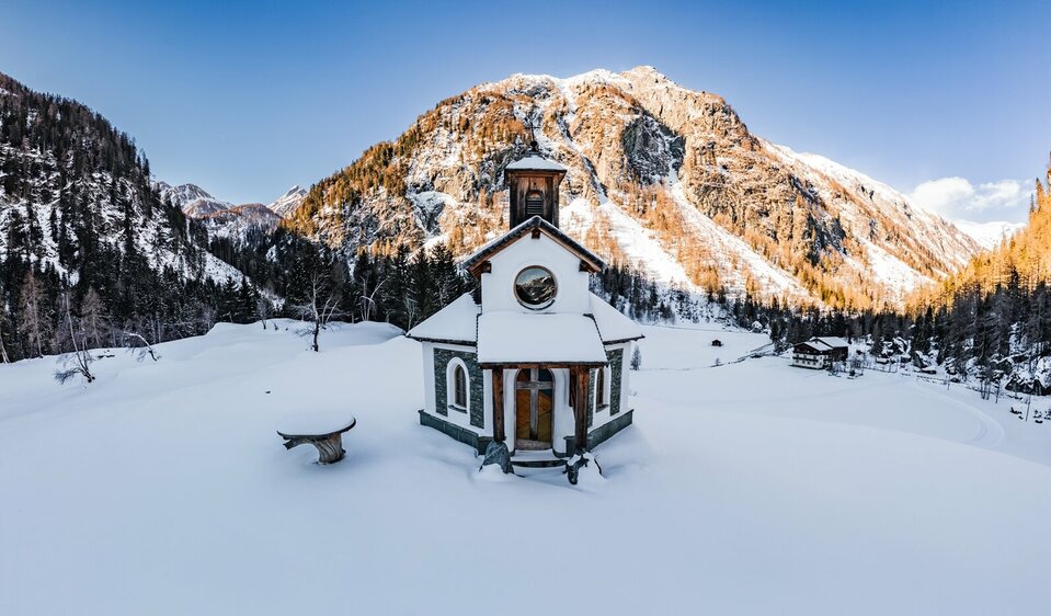 Frontansicht der schönen aus Holz und Stein gebauten Kapelle in Prägraten im Schnee. Im Hintergrund erhebt sich ein von der Sonne angestrahlter Berg.
