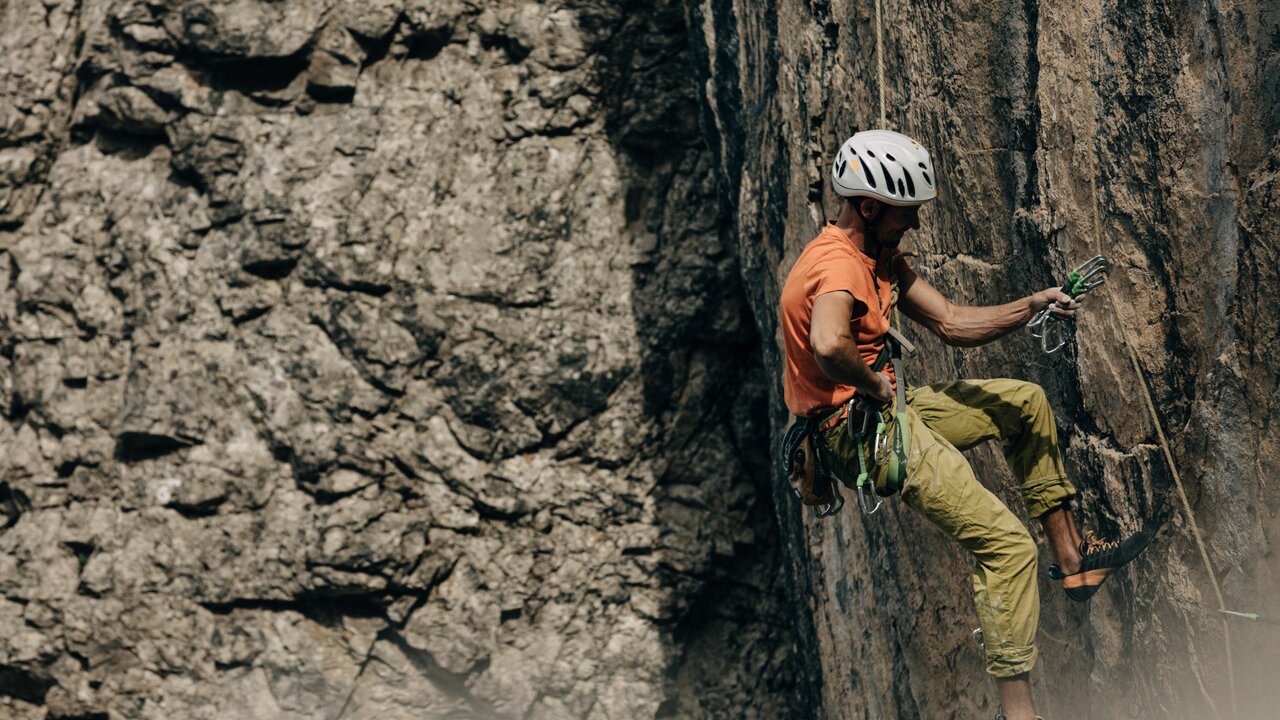 Laserz Klettersteig Ein Kletterer mit orangenem Shirt und weißem Helm, hängt an einer steilen Felswand am Laserz Klettersteig.