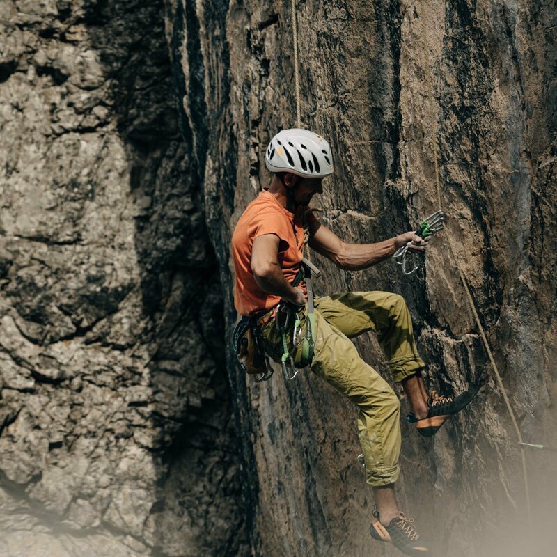 Ein Kletterer mit orangenem Shirt und weißem Helm, hängt an einer steilen Felswand am Laserz Klettersteig.