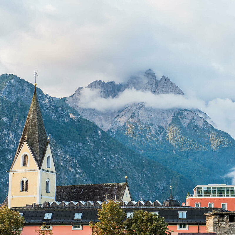Blick auf die Hausdächer und den Kirchturm in Lienz mit beeindruckender Bergkulisse im Hintergrund.