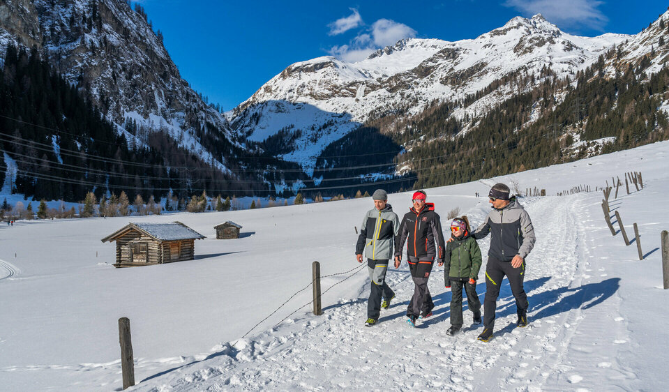 Eine Familie, bestehend aus Mutter, Vater und zwei Kindern, wandern lachend in tief verschneiter Winterkulisse über einen Winterwanderweg.