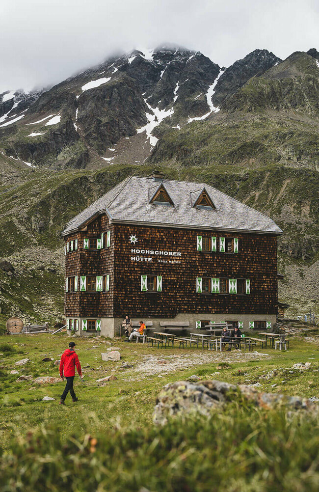Eine Person mit roter Jacke geht kurz vor der Hochschober Hütte. Links steht eine grüne Alpenvereins-Flagge.