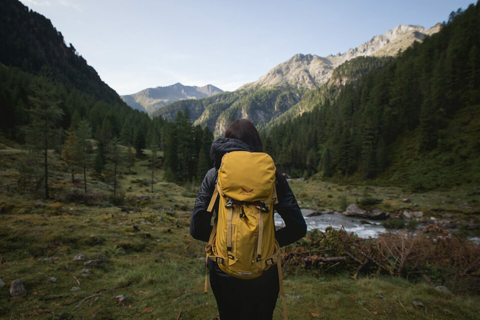 Wandern in Osttirol Wanderin mit gelbem Rucksack auf einer Lichtung in morgendlicher Stimmung kurz vor einem kleinen Gebirgsbach. Im Hintergrund strahlt die Morgensonne schon einen Bergrücken an.