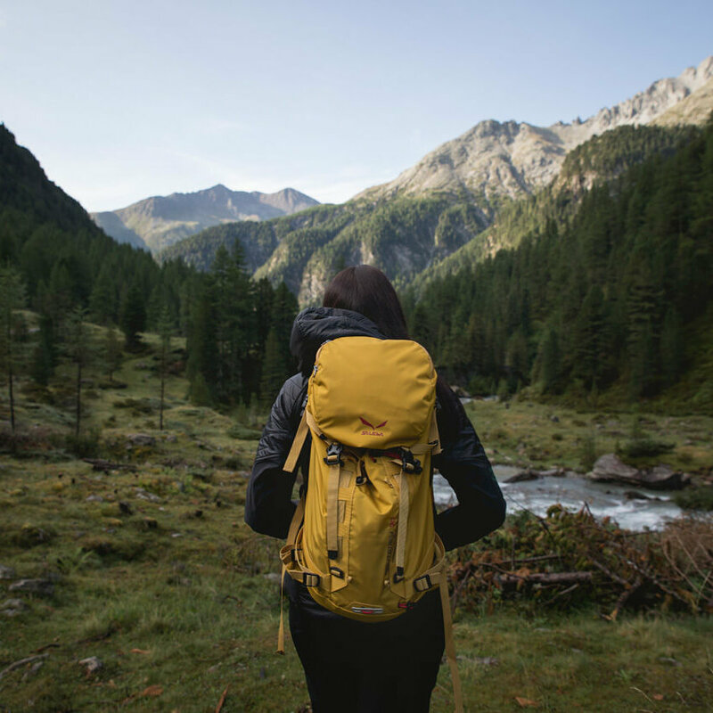 Wandern in Osttirol Wanderin mit gelbem Rucksack auf einer Lichtung in morgendlicher Stimmung kurz vor einem kleinen Gebirgsbach. Im Hintergrund strahlt die Morgensonne schon einen Bergrücken an.
