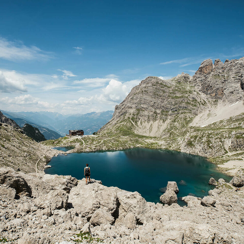 Eine Wandererin blickt auf die Karlsbader Huette und den davor liegenden Laserzsee bei Traumwetter.