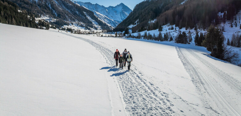 Eine Familie, bestehend aus zwei Elternteilen und zwei Kindern, wandert inmitten verschneiter Bergwelt. Die Sonne strahlt am wolkenlosen, blauen Himmel.