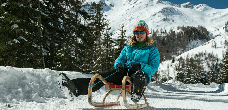 Eine Frau mit blauer Jacke und bunter Mütze rodelt mit einem Schlitten ins Tal.