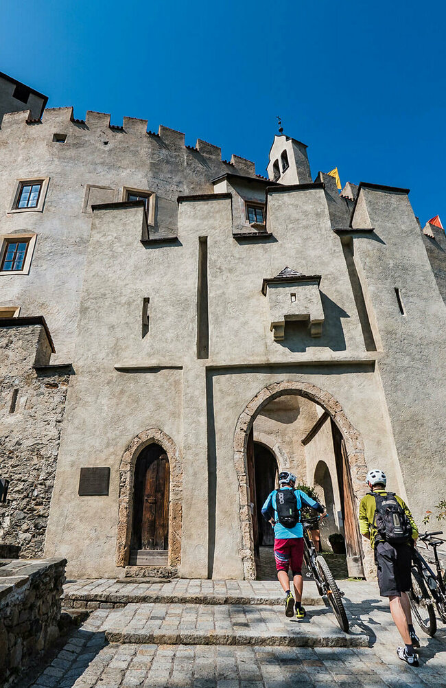 Zwei Mountainbiker, die ihre Mountainbikes über einen breiten, gepflasterten Zugang zu einem alten, Schlosseingang über einen niederen Treppenabsatz schieben. Der linke Biker trägt eine weinrote, knielange Hose und eine hellblaue Radjacke, der zweite Radfahrer eine graue, knielange Hose und eine moosgrüne Jacke. Beide tragen auch einen weißen Radhelm sowie dunkle Rucksäcke. Die Schlossmauern sind vom Sonnenlicht angestrahlt und darüber blitzt blauer Himmel.