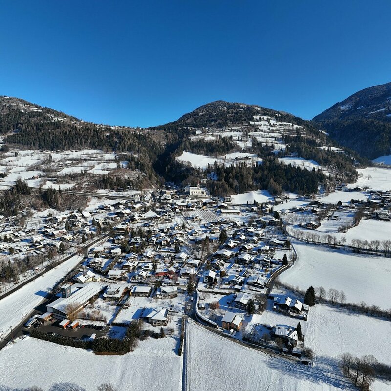 L'immagine mostra il paesaggio invernale del paese di Dölsach, situato su una piccola altura. I tetti, i campi, i prati e i pendii sono interamente ricoperti di neve. Il paese è circondato da dolci colline boscose e catene montuose. Il cielo è sereno e splende il sole.