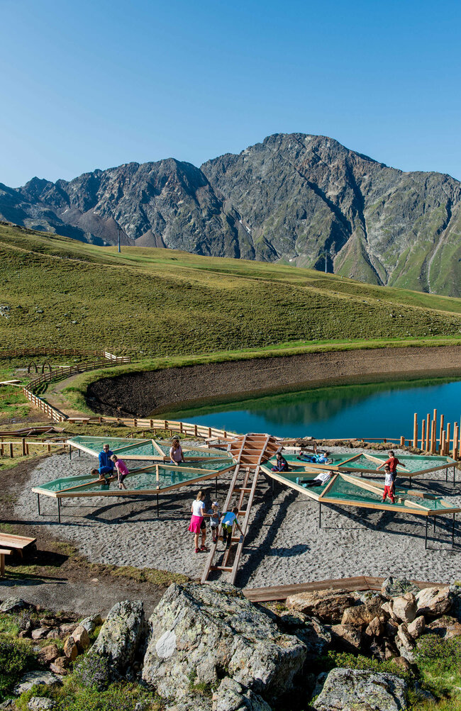 Wassermythos Ochsenlacke St. Jakob Brunnalm Zu sehen sind mehrere Kinder die auf einem Spielgerät, welches einer überdimensional-großen Libelle ähnlich sieht, beim Wassermythos Ochsenlacke im Skizentrum St. Jakob i. D. spielen. Das sonnige Wetter lässt den sichtbaren Teil des Ufers und die dahinterliegende Bergwelt in einem warmen Licht erstrahlen.