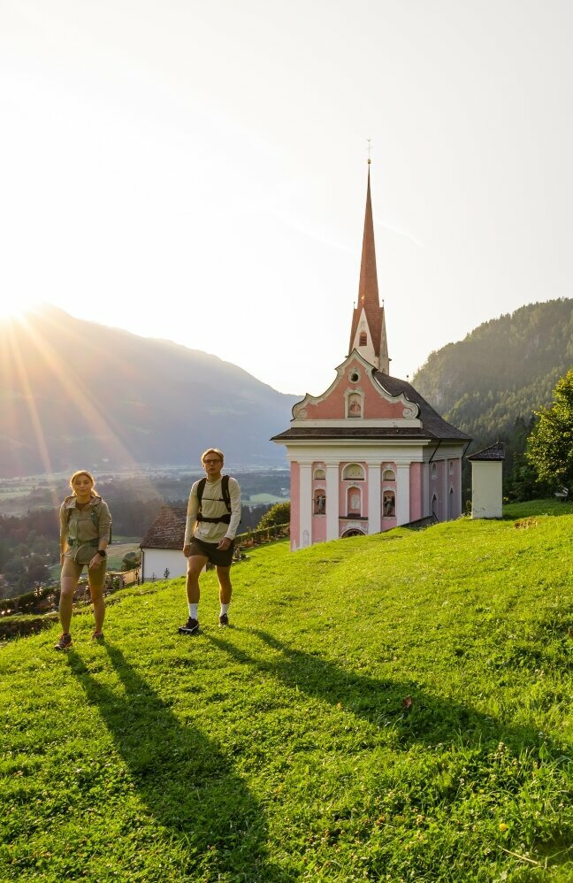 Etappe 1 - Pfarrkirche St. Ulrich in Lavant Ein Pärchen wandert auf dem Hoch und Heilig Weitwanderweg vor der Pfarrkirche St. Ulrich in Lavant im Sonnenuntergang.