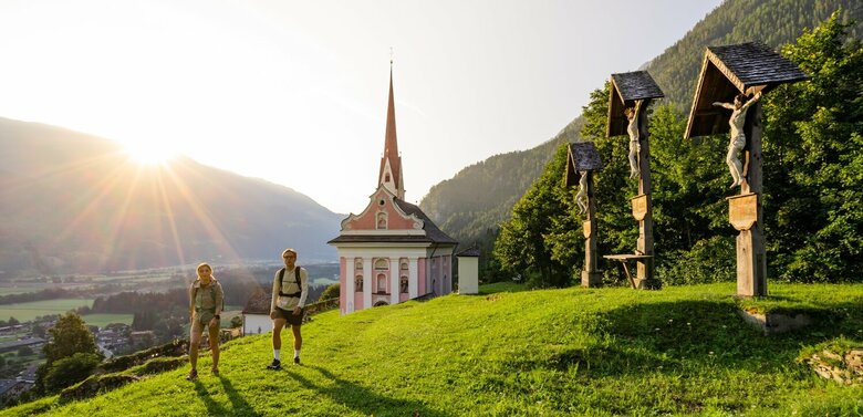 Ein Pärchen wandert auf dem Hoch und Heilig Weitwanderweg vor der Pfarrkirche St. Ulrich in Lavant im Sonnenuntergang.