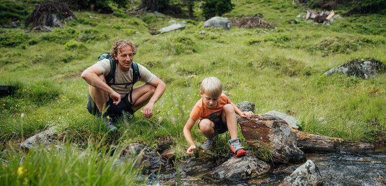 Ein Vater und ein Sohn sitzen an einem Gebirgsbach und lassen ein aus Naturmaterialien selbstgebautes Schiff im Wasser entlang gleiten.
