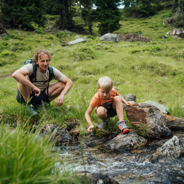 Ein Vater und ein Sohn sitzen an einem Gebirgsbach und lassen ein aus Naturmaterialien selbstgebautes Schiff im Wasser entlang gleiten.