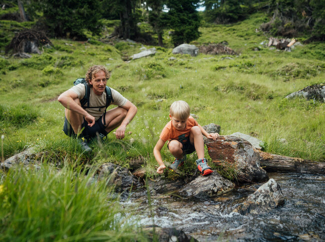 Ein Vater und ein Sohn sitzen an einem Gebirgsbach und lassen ein aus Naturmaterialien selbstgebautes Schiff im Wasser entlang gleiten.
