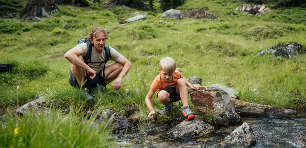 Ein Vater und ein Sohn sitzen an einem Gebirgsbach und lassen ein aus Naturmaterialien selbstgebautes Schiff im Wasser entlang gleiten.