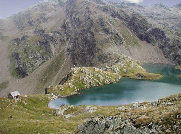Der Blick von etwas oberhalb auf den Geigensee in Hopfgarten i. D., der seinen Namen seiner geigenförmigen Form zu verdanken hat. Das blaue Wasser hebt sich eindeutig von der grün-grauen, felsigen Umgebung ab. Links vom See steht eine kleine Hütte.