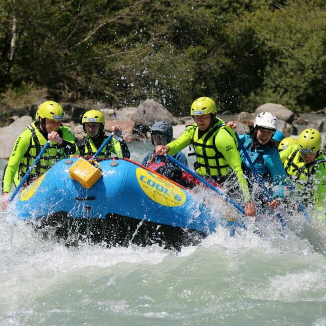 Eine Gruppe von Menschen beim Wildwasser-Rafting auf einem Fluss. Sie sitzen gemeinsam in einem blauen Schlauchboot. Sie tragen Schutzkleidung wie Helme und Schwimmwesten in auffälligen Farben. 