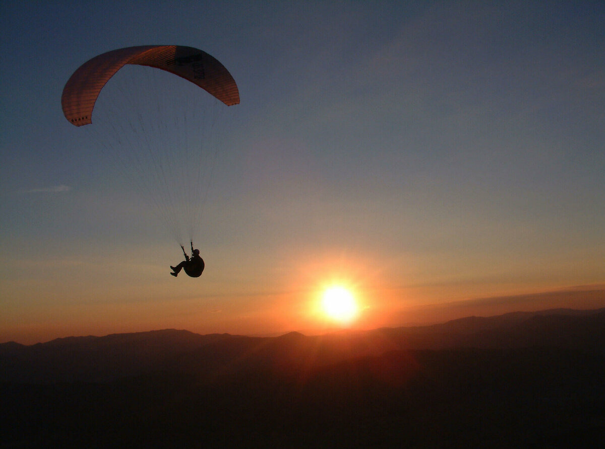Paragleiter am abendlichen Himmel, dahinter Sonne, die bald am Horizont verschwindet.