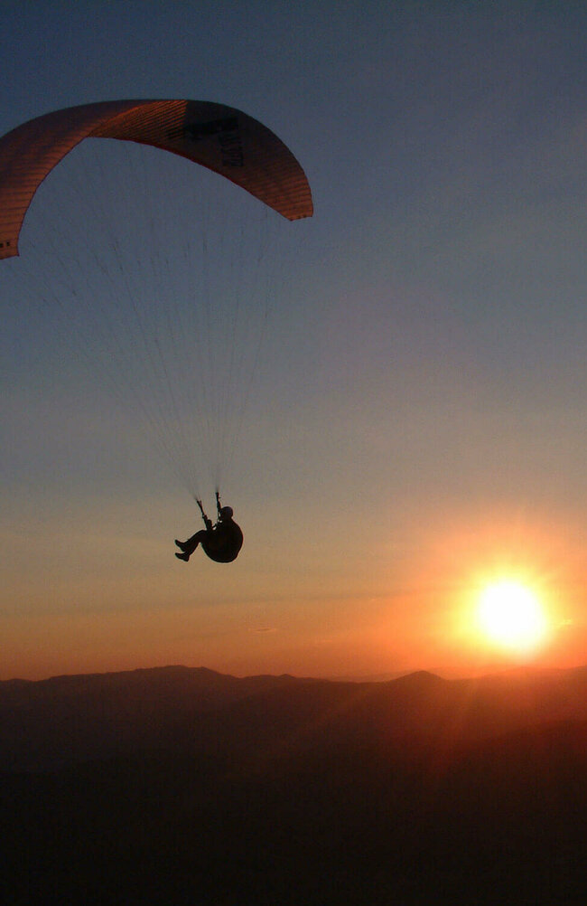 Paragleiter am abendlichen Himmel, dahinter Sonne, die bald am Horizont verschwindet.