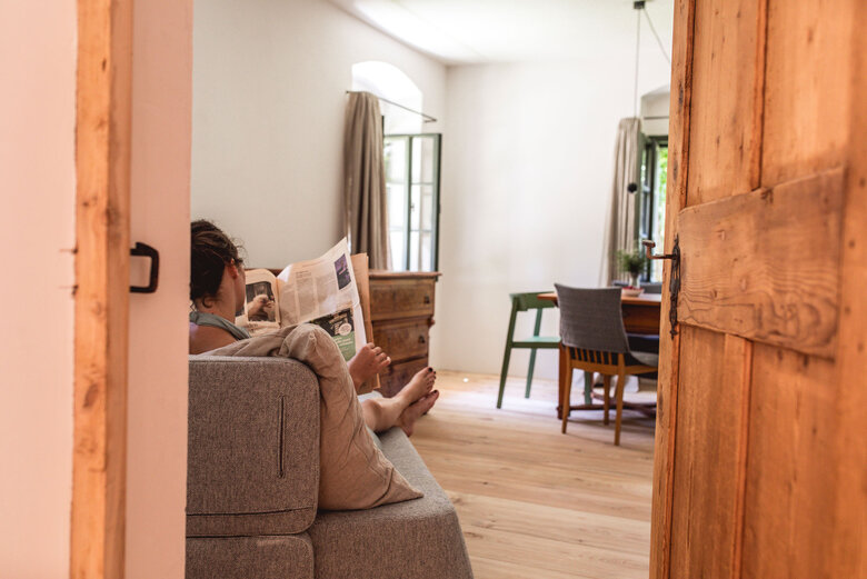 Entspannung im Iselhof Eine Frau entspannt in ihrer Unterkunft auf einem Sofa und liest Zeitung.