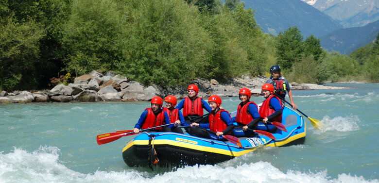 Ein Raftingboot mit mehreren Personen in Neoprenanzügen mit roten Schwimmwesten und roten Helmen auf der Isel.