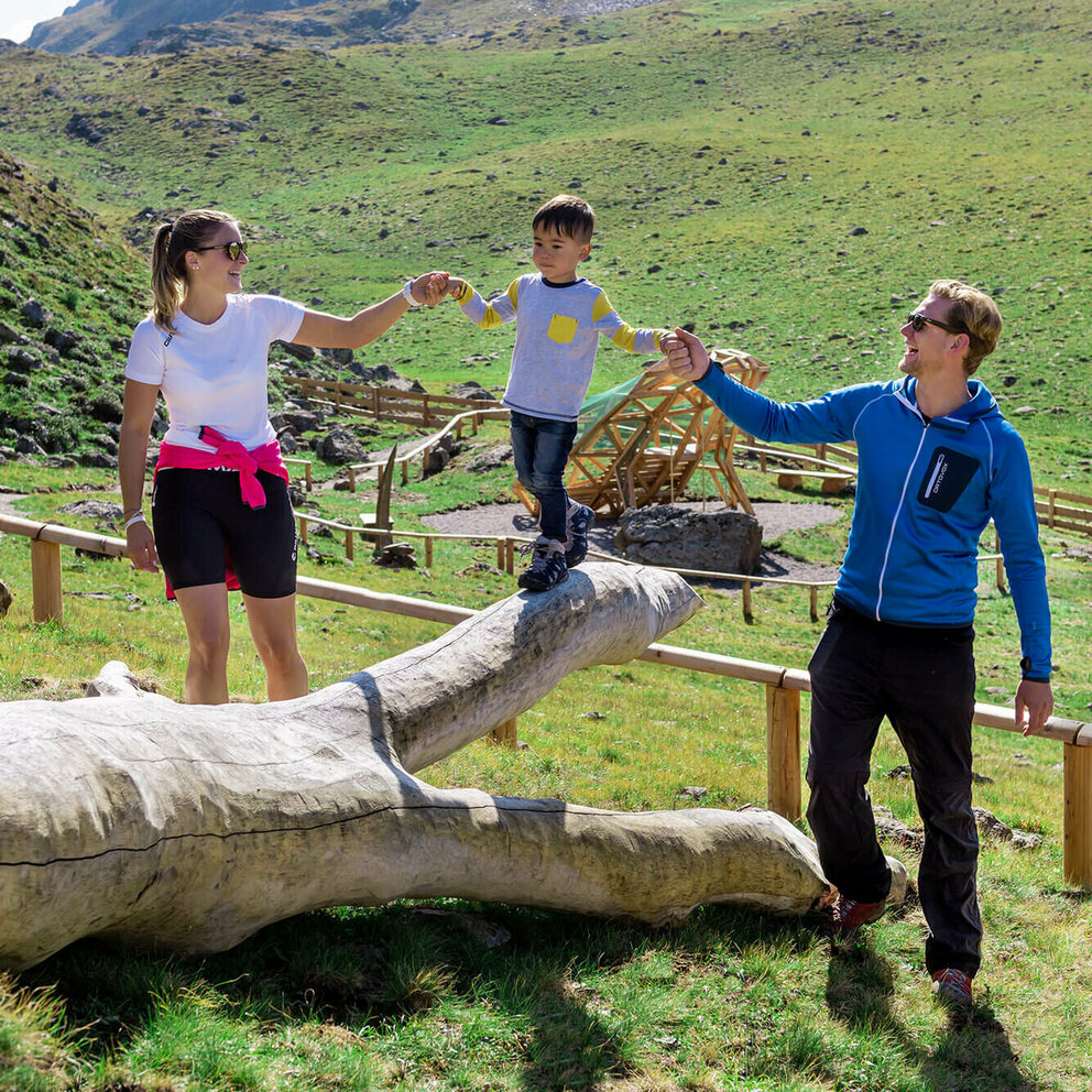 Eine Familie genießt die Zeit beim Wassermythos Ochsenlacke im Skizentrum St. Jakob i. D.. Ein Kind balanciert auf einem Spielgerät während er links und rechts von seinen Eltern dabei geführt wird. Die Sonne taucht die Umgebung in ein warmes Licht.
