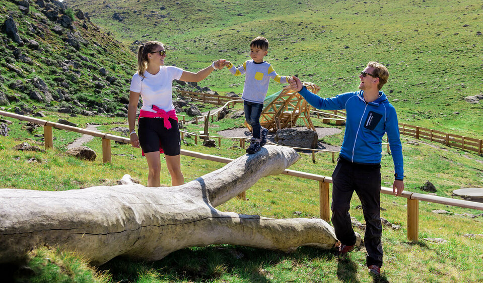 Eine Familie genießt die Zeit beim Wassermythos Ochsenlacke im Skizentrum St. Jakob i. D.. Ein Kind balanciert auf einem Spielgerät während er links und rechts von seinen Eltern dabei geführt wird. Die Sonne taucht die Umgebung in ein warmes Licht.