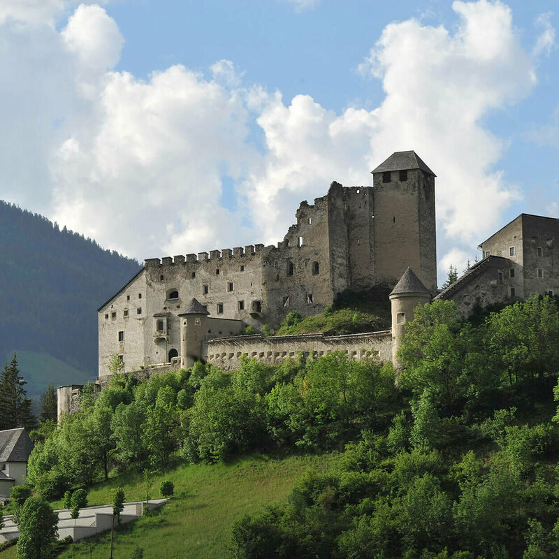 Majestätischer Sommerblick aus dem Tal auf die Burg Heinfels, eingebettet in grüne Laubbäume im satten Sommerkleid