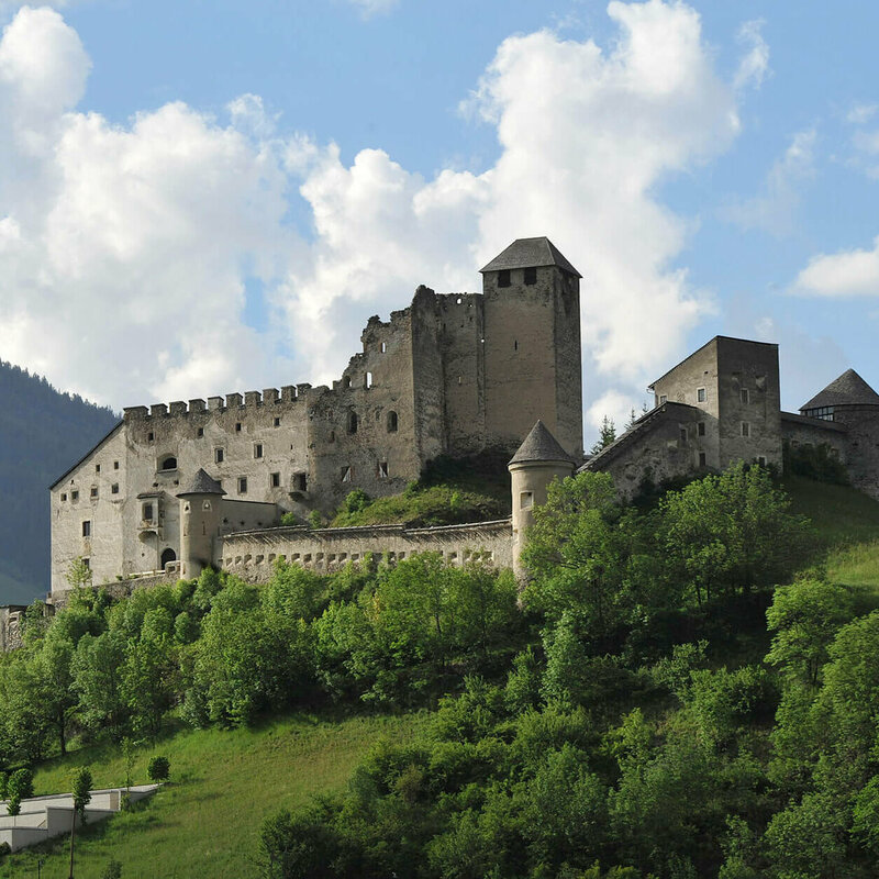 Burg Heinfels Majestätischer Sommerblick aus dem Tal auf die Burg Heinfels, eingebettet in grüne Laubbäume im satten Sommerkleid