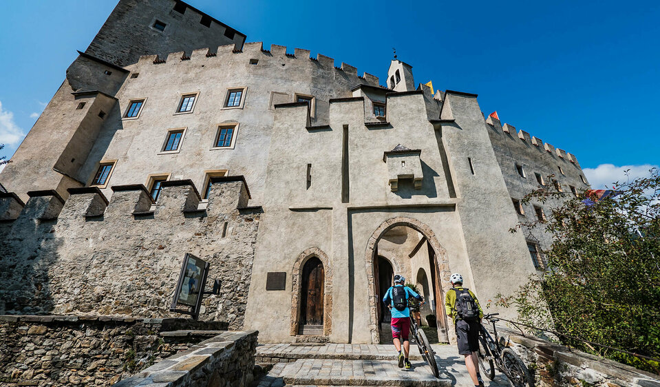 Schloss Bruck Zwei Radfahrer schieben ihre Fahrräder durch einen steinernen Eingangsbogen in das Schloss Bruck in Lienz.