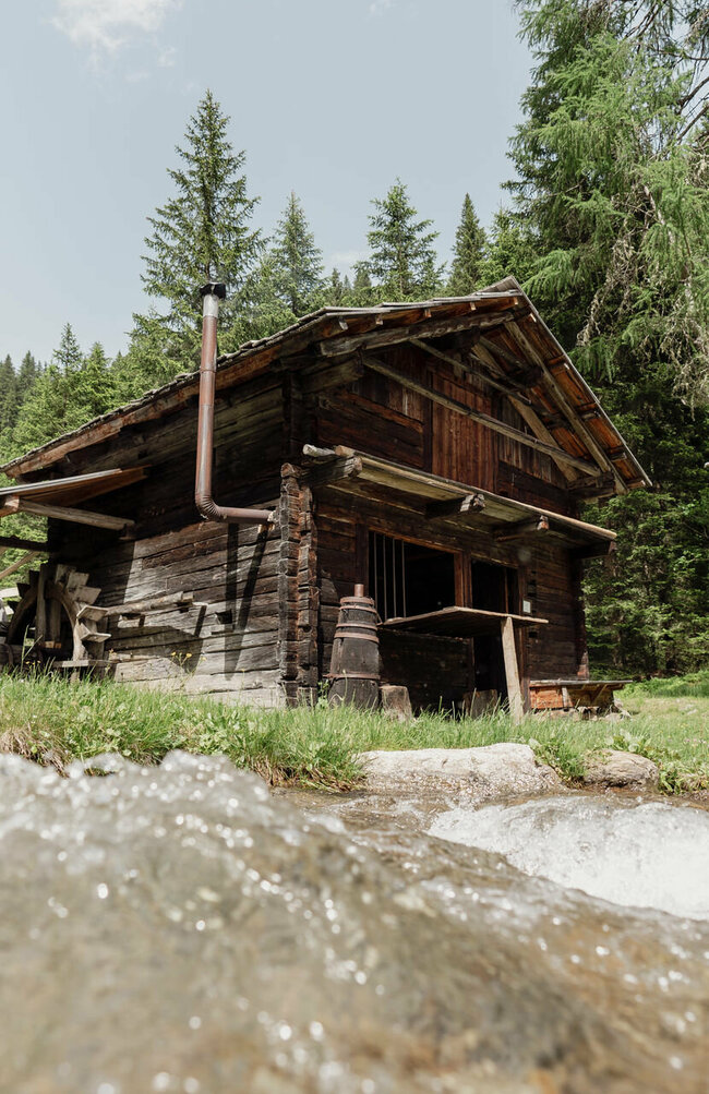 Sommeraufnahme der Mühle im Freilichtmuseum Alpines Leben in Innervillgraten, im Vordergrund fließt ein klarer Bach