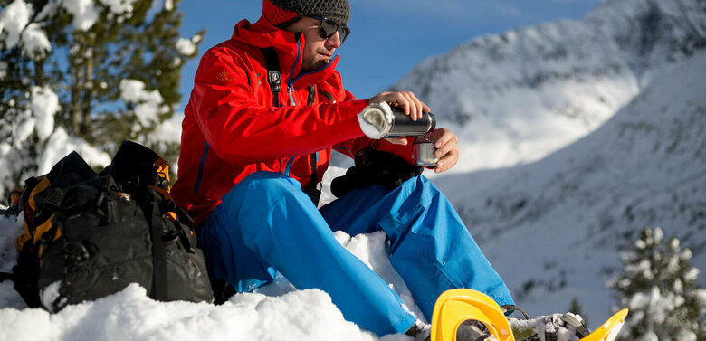 Ein Mann mit roter Jacke und Mütze macht Rast und schenkt sich ein warmes Getränk ein. Er ist am Schneeschuhwandern im Defereggental.