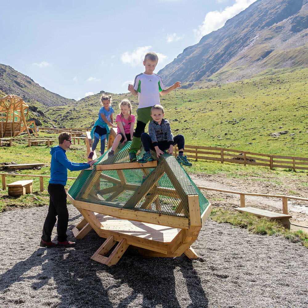 Spielende Kinder beim Wassermythos Ochsenlacke im Skizentrum St. Jakob i. D.. Sie klettern auf einem Spielgerät, was ihnen sichtlich viel Spaß bereitet. Die Sonne lässt die Umgebung und die dahinterliegende Bergwelt in einem warmen Licht erstrahlen.