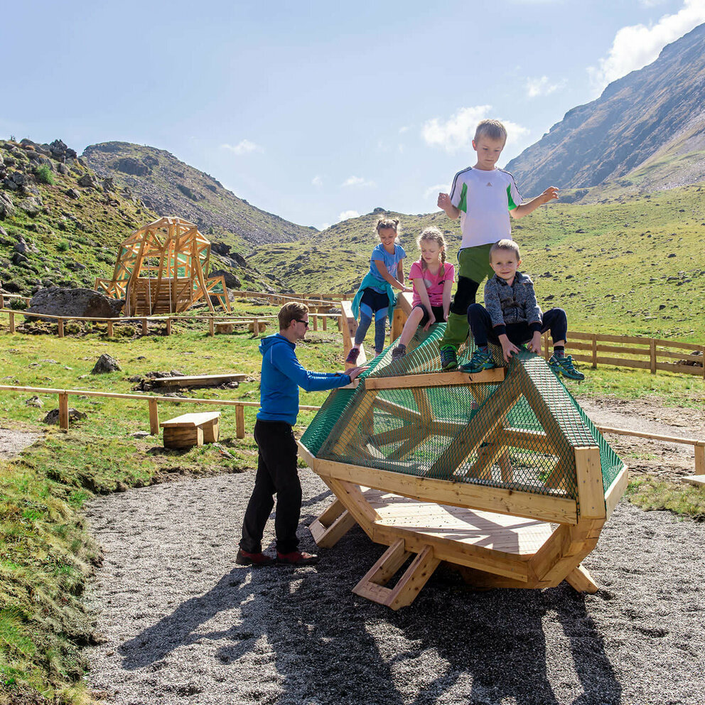 Spielende Kinder beim Wassermythos Ochsenlacke im Skizentrum St. Jakob i. D.. Sie klettern auf einem Spielgerät, was ihnen sichtlich viel Spaß bereitet. Die Sonne lässt die Umgebung und die dahinterliegende Bergwelt in einem warmen Licht erstrahlen.