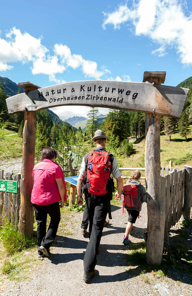 Eine Familie in Wanderkleidung geht durch das aus Holz gebaute Eingangstor am Oberhauser Zirbenwald.