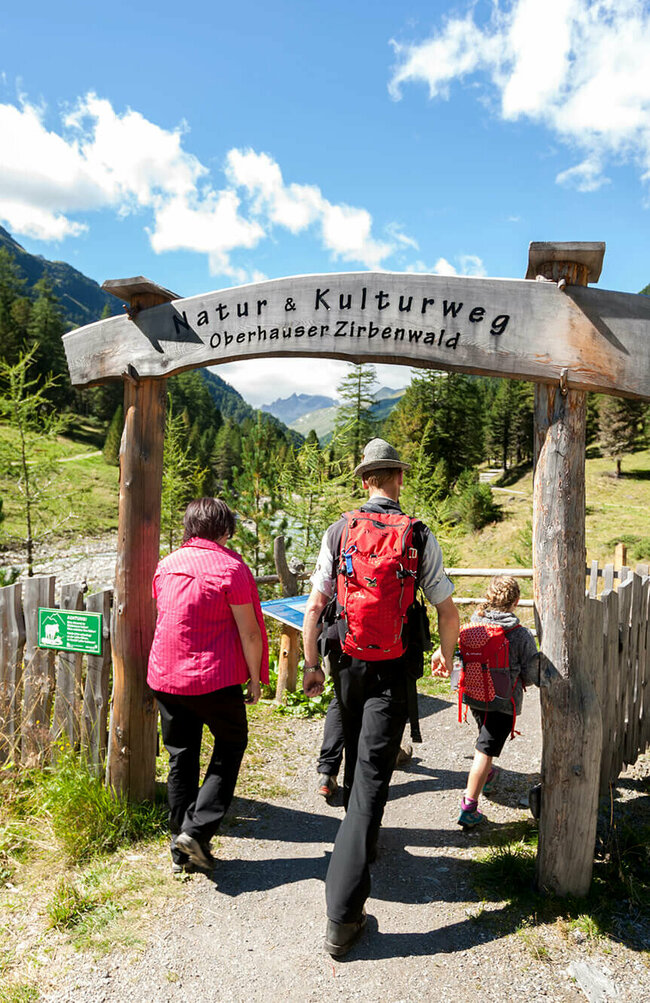 Eine Familie in Wanderkleidung geht durch das aus Holz gebaute Eingangstor am Oberhauser Zirbenwald.