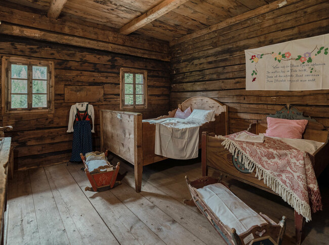 Schalfzimmer im Freilichtmuseum Alpines Leben In einem alten Baunerhaus stehen zwei Doppelbetten und zwei Wiegen. Ein traditionelles Trachtengewand wird stolz ausgestellt, während ein kunstvolles Wandtuch die Wand ziert.