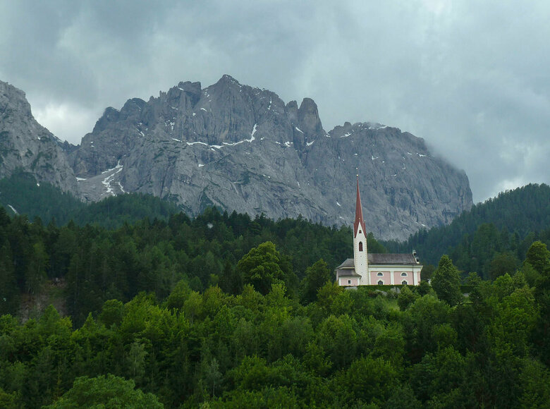 Die Kirche St. Ulrich in Lavant ragt aus dem dicht bewaldeten Hügel hervor. Im Hintergrund sieht man die Ausläufer der Lienzer Dolomiten. 