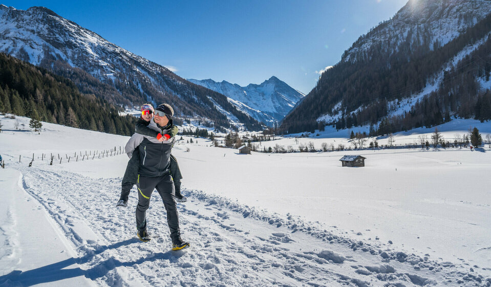 Ein Vater trägt seine Tochter in verschneiter Berglandschaft auf dem Rücken. Im Hintergrund sieht man verschneite Berggipfel und dunkelgrüne Wälder im strahlenden Sonnenlicht.