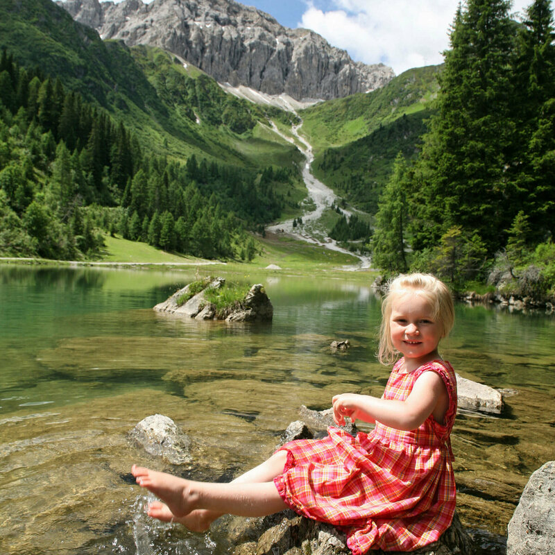 Der Klapfsee - Magnet für Erholungssuchende Mädchen in rot-kariertem Sommerkleidchen auf einem Stein am Ufer des Klapfsees sitzend, im Hintergrund rechts mit Nadelbäumen und Felsen begrenztes Seeufer und ganz im Hintergrund das Bergmassiv der Porze aus Kalkgestein. Darüber leicht bewölkter Himmel.
