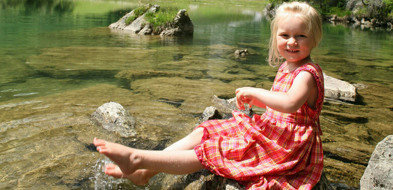Mädchen in rot-kariertem Sommerkleidchen auf einem Stein am Ufer des Klapfsees sitzend, im Hintergrund rechts mit Nadelbäumen und Felsen begrenztes Seeufer und ganz im Hintergrund das Bergmassiv der Porze aus Kalkgestein. Darüber leicht bewölkter Himmel.
